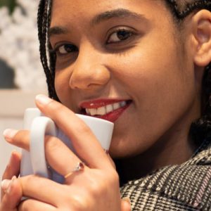 Image of a pretty lady, with brown eyes and red lipstick, smiling, while cradling a cup that she is about to drink from.