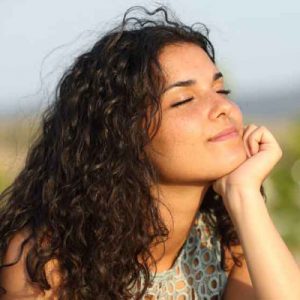 Female with long wavy brown hair, resting her chin on her hand, smiling with her eyes closed. Illustrating that Meditation helps us to feel relaxed and rejuvenated