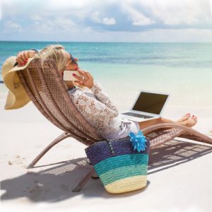 Female in her 50s, relaxed me some chair on a tropical beach. Holding a telephone in one hand, a sun hat in the oven, and a laptop resting on her legs.