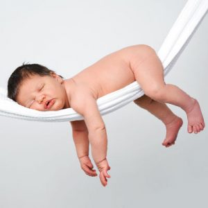 image of a baby with short dark hair, laying face down on a white hammock, with their arms dangling over this side.