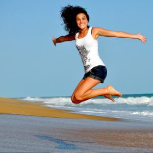 Image of a happy female leaping up in the air, from a sandy beach, what is the waves are receiving back to the sea?. Her arms are stretched outwards, and she is smiling and looking very happy